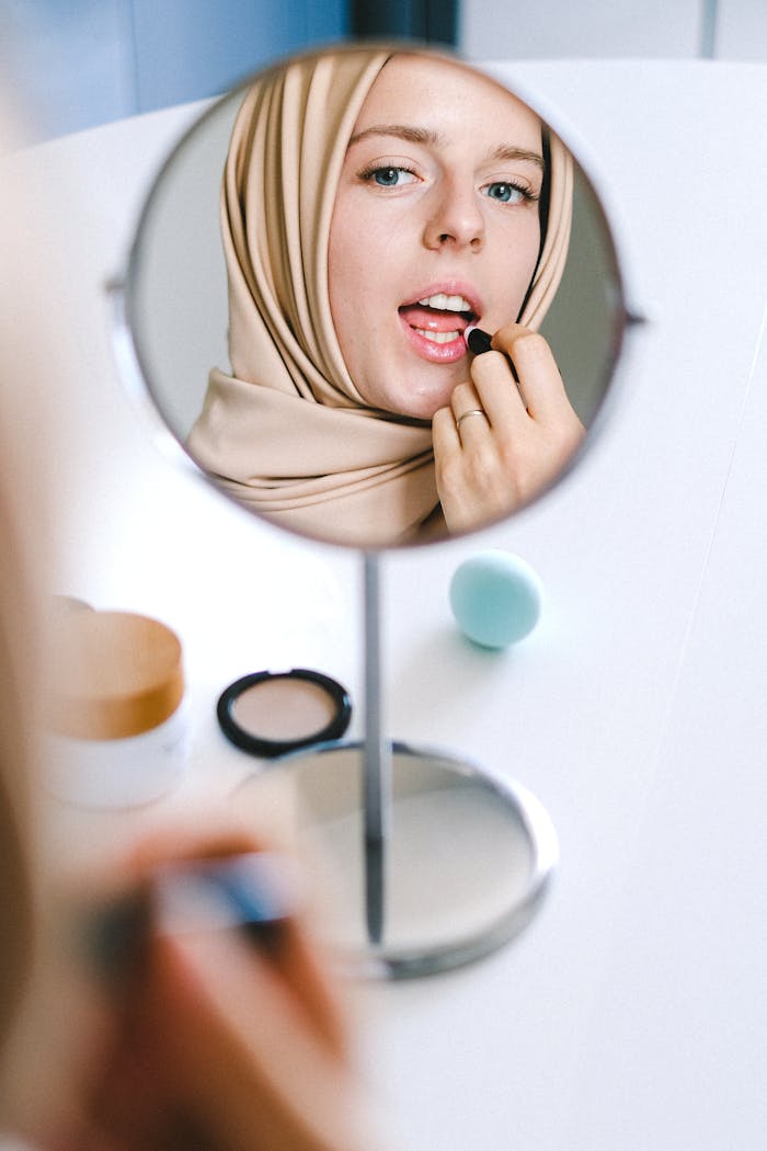 A woman in a hijab applying lipstick, captured in a mirror reflection, with beauty products on a table.
