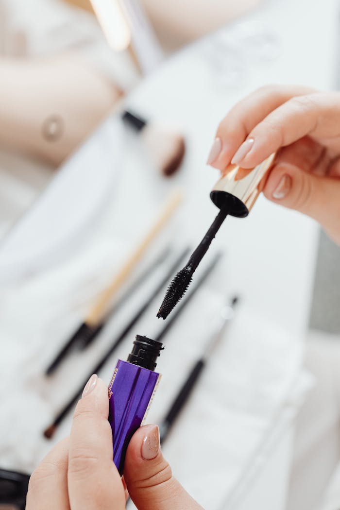 High-quality close-up of a mascara brush held in hand, surrounded by various makeup tools.